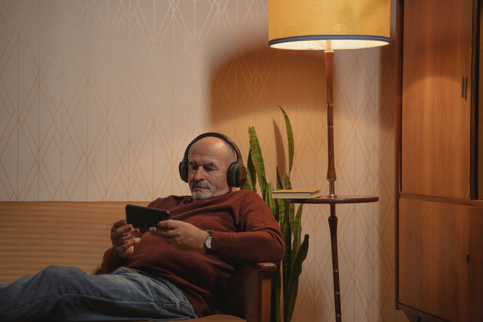Älterer Herr abends auf dem Sofa unter einer Stehlampe, mit Kopfhörern und Smartphone